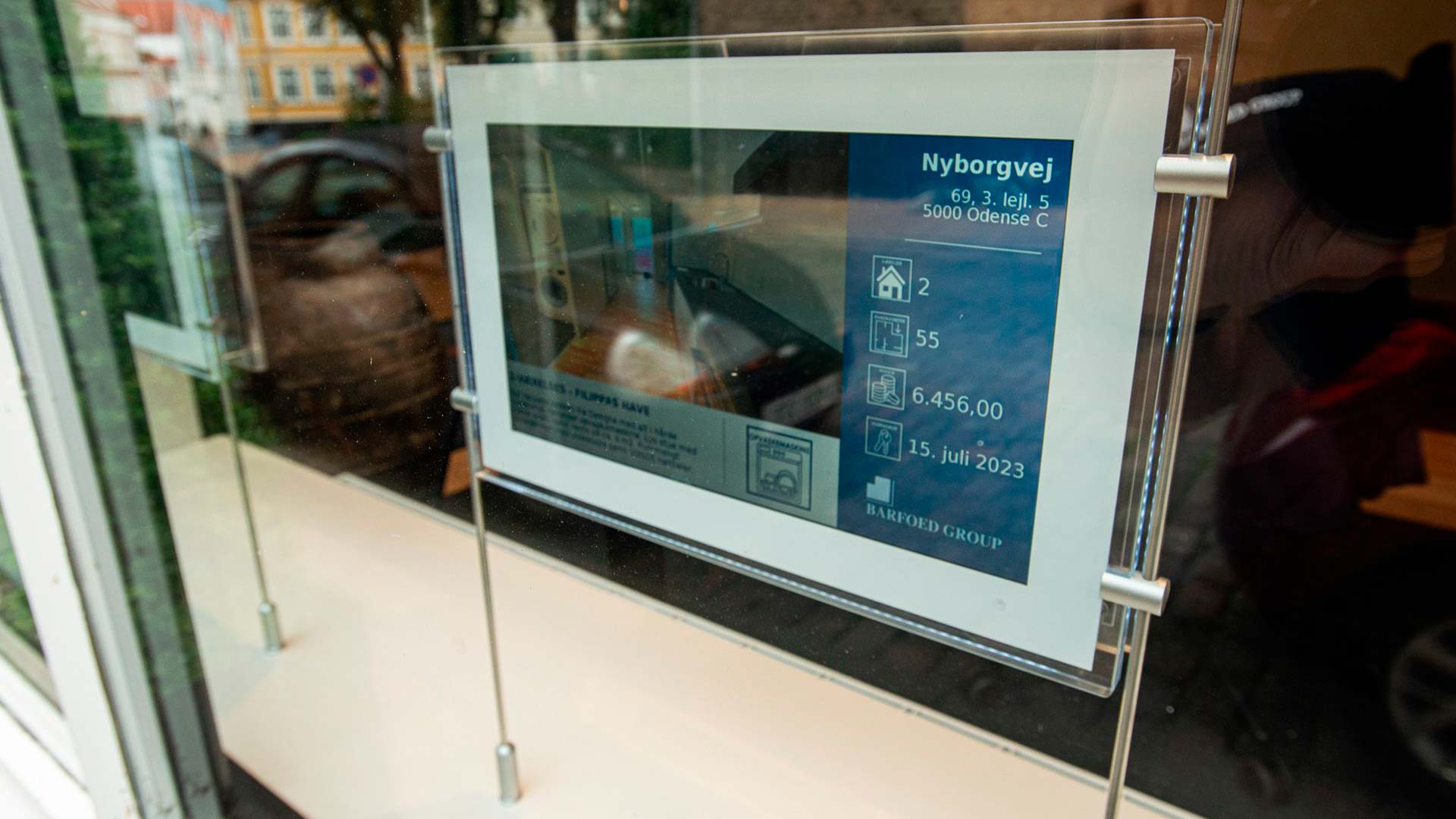 Digitalt mæglerskilt i vinduet hos ejendomsmægler, viser ejendomsdetaljer tydeligt.