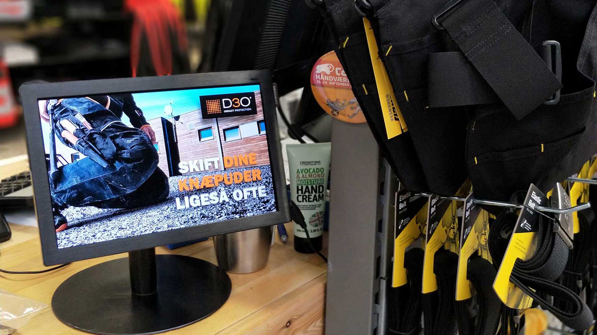 Digitalt butiksdisplay på 10 tommer installeret ved kassen hos Carl Ras i Herlev, der viser Snickers Workwear kampagne og påminder kunder om knæbeskyttere